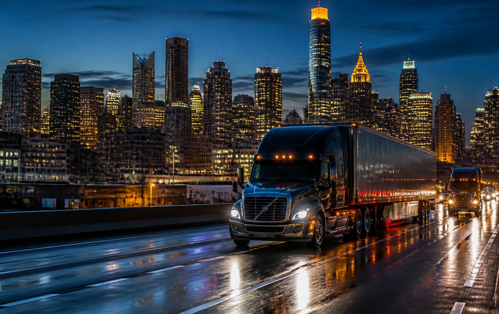 Tractor-trailers moving along a wet highway at night with a lit city skyline behind them